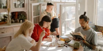 Four young adults study together at a sunlit table covered with books and papers, while another person reads by an open window in the background. The atmosphere is relaxed and focused.