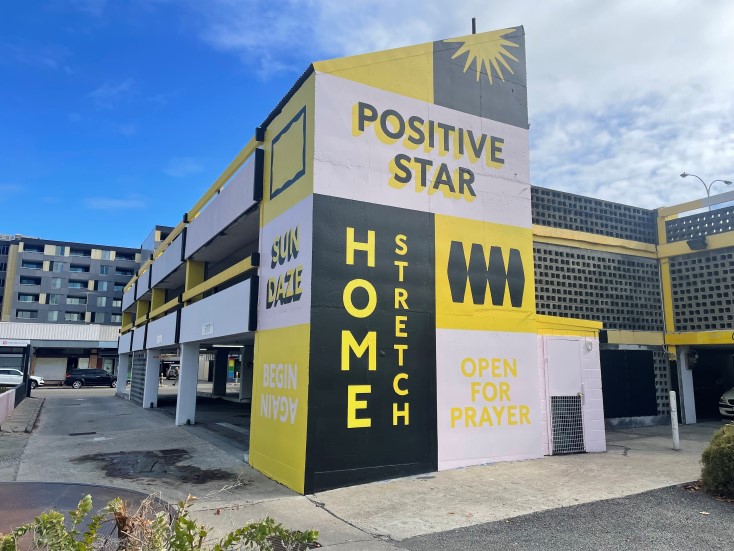 A two-story parking garage painted with bold yellow, black, and white blocks. Words like “POSITIVE STAR,” “SUN DAZE,” “HOME STRETCH,” “BEGIN NOW,” and “OPEN FOR PRAYER” are displayed on the exterior.