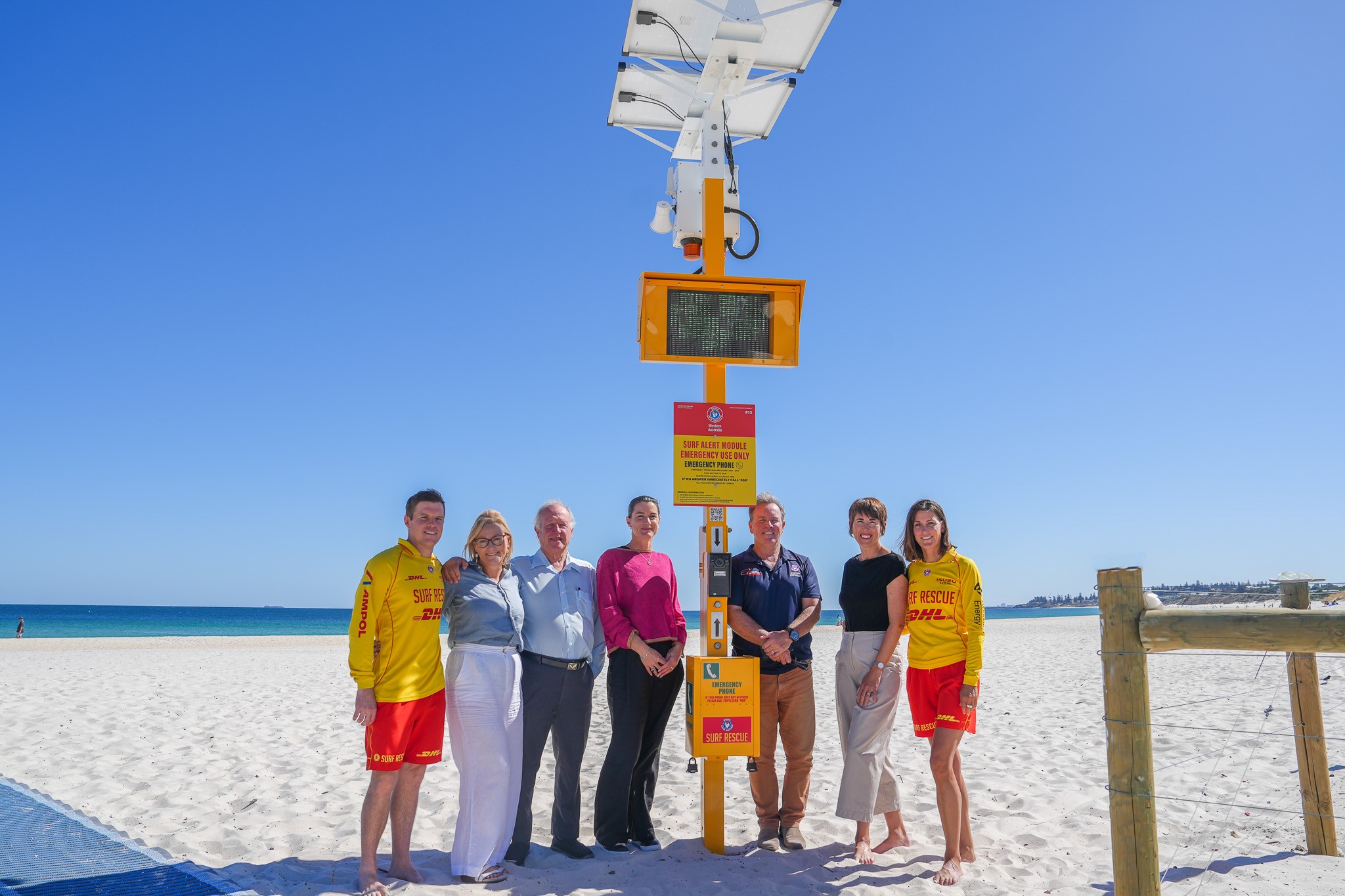 Six people, including two lifeguards in yellow and red uniforms, stand on a sandy beach next to a solar-powered emergency information sign under a clear blue sky.