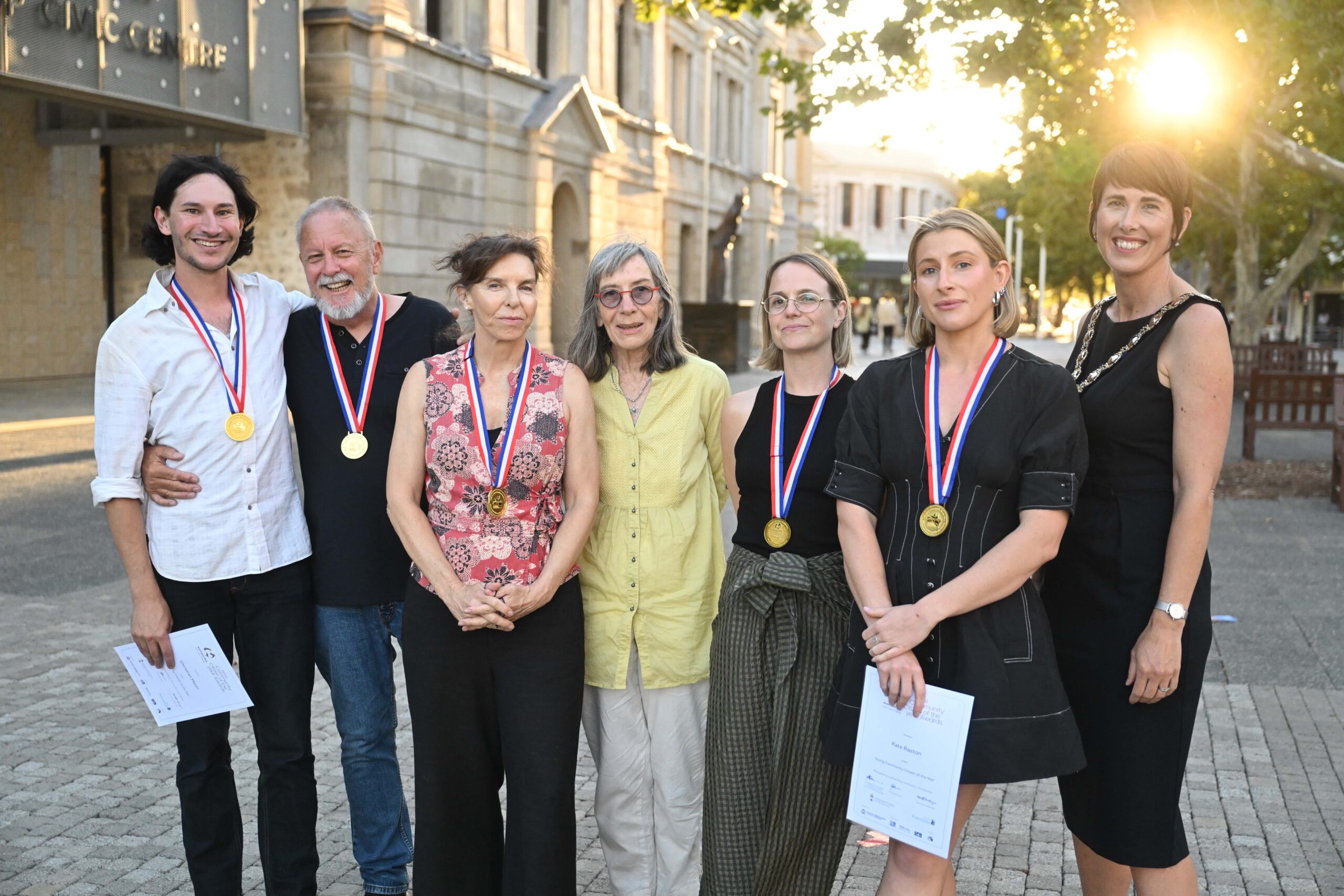 A group of seven people stand outdoors, some wearing gold medals around their necks and holding certificates. They are smiling and posing for the photo on a sunny day near a stone building and trees.