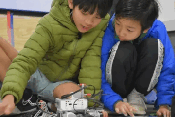 Two children sit on the floor, closely examining and interacting with a small robot made of plastic and metal parts, appearing focused and engaged in a STEM activity.