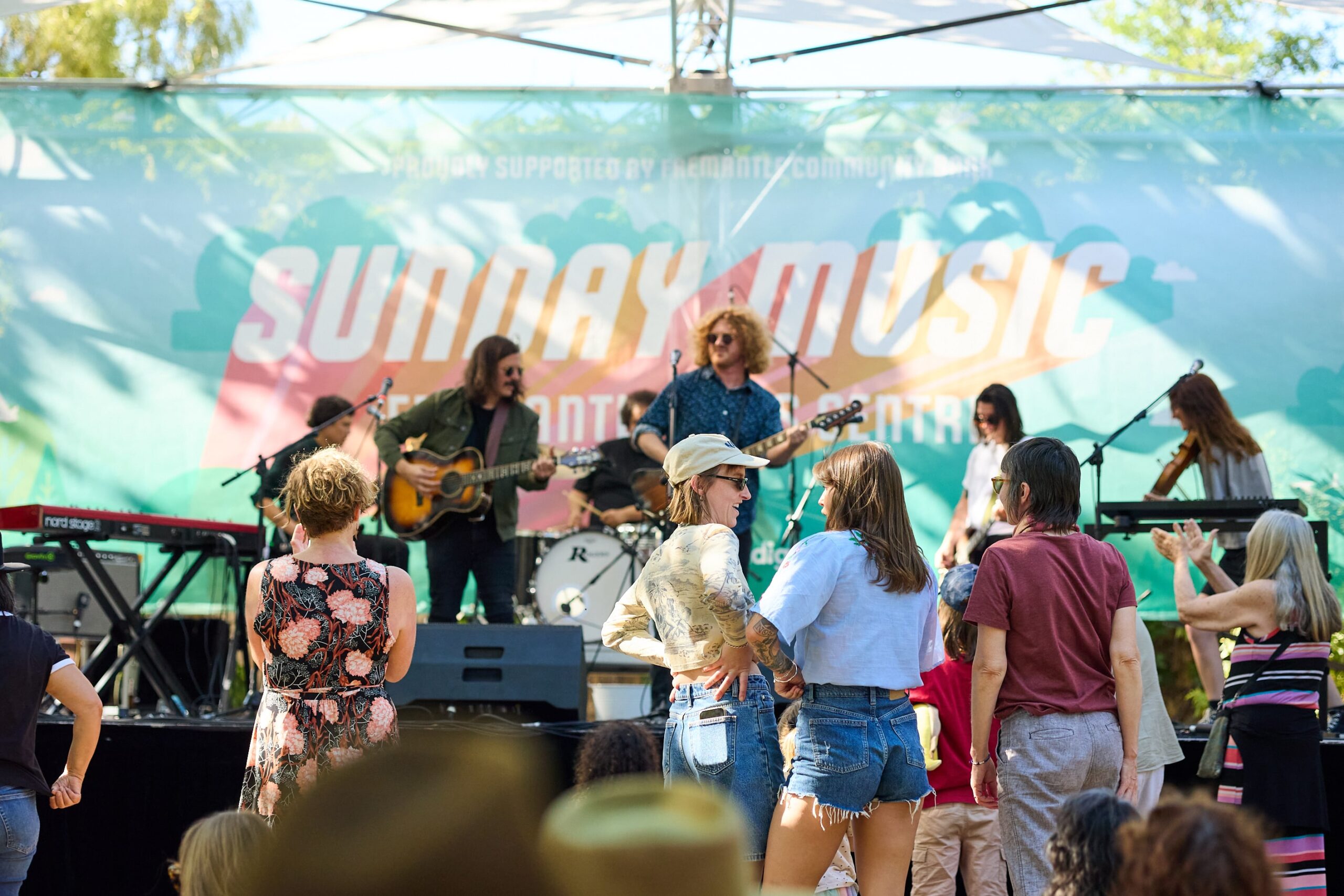 People enjoy live music outdoors, with a band playing on stage under a “Sunday Music” banner. Audience members stand, dance, and watch in casual clothing, framed by sunlight and trees.