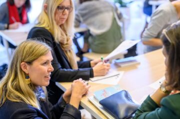 Three women sit at a table, engaged in conversation and writing on papers. One woman in the foreground is speaking, while another writes notes. A purse and book are on the table. Other people are blurred in the background.