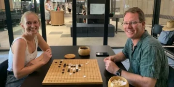 Two people smiling while playing a game of Go at a black table indoors. Black and white stones are on the board, with bowls of pieces nearby. Other people and tables can be seen in the background.