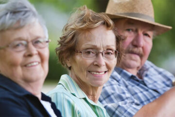 Three older adults sit closely together outdoors, smiling at the camera. The woman in the center wears glasses and a striped shirt, while the man beside her wears a straw hat. The background is green and blurred.