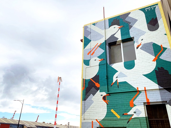 A building exterior features a colorful mural of stylized seagulls with orange beaks and legs, set against geometric blue, green, and white shapes. The sky is cloudy and industrial structures are visible in the background.