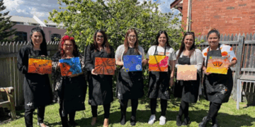 Seven women stand in a row outside, each holding a colorful painting. They wear black aprons and smile at the camera. A tree, fence, and brick building are visible in the background.