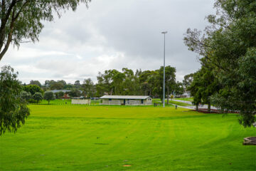 A wide, green grassy field with a small white building and tall light pole in the background, surrounded by trees under a cloudy sky.