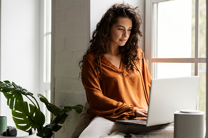 A woman with long curly hair sits by a window, wearing an orange blouse and using a laptop. Natural light comes through the window, and there is a green potted plant nearby.