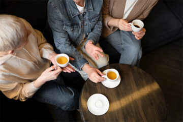Three people sitting around a wooden table, holding white teacups with tea, engaged in conversation. Only their hands and partial torsos are visible, showing casual, comfortable clothing.