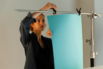 A person attaches a large sheet of light blue paper to a horizontal metal rod using clamps, possibly preparing a backdrop for a photoshoot in a well-lit indoor setting.