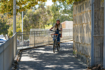 A woman in a helmet rides a bicycle with a front basket along a paved path between a metal fence and a stone wall.