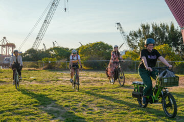 Four people wearing helmets ride bicycles across a grassy park with industrial cranes in the background.