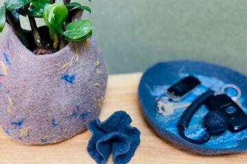 A small plant in a soft, multicolored felt pot sits on a wooden surface next to a round blue felt tray holding keys and a key fob, with a dark blue felt object nearby.