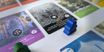 A close-up of colorful board game cards and blue and green meeple pieces on a white surface, featuring intricate patterns and symbols on the cards.