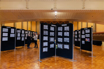 Exhibition hall with parquet floor displaying multiple black panels covered in black-and-white sketches and drawings. A few people are examining the artwork. The hall has ornate details and warm lighting.
