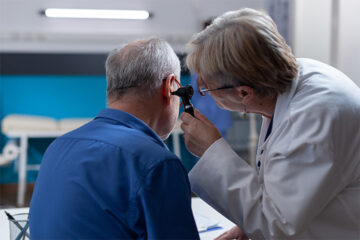 A doctor examines an older mans ear using an otoscope in a medical office. The man is seated and wearing a blue shirt; the doctor is focused on his ear, checking for any issues.