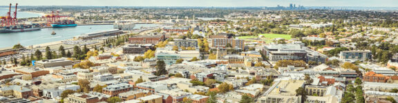Aerial view of a coastal city with a river, industrial docks, and a mix of residential and commercial buildings; green parks and a distant city skyline are visible under a clear sky.