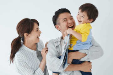 A smiling man holds a laughing young boy in his arms while a woman stands beside them, also smiling. The family appears happy and engaged together against a plain white background.