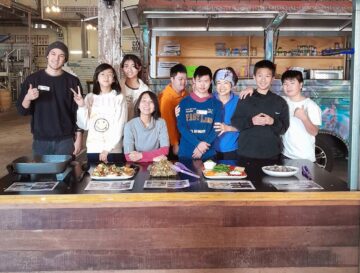 A group of ten people stand smiling together behind a table with plates of food, inside an indoor space with a food truck and shelves in the background. Some people are making peace signs.