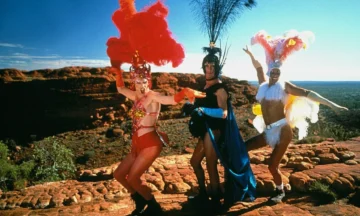 Three people in colorful, flamboyant costumes with feathers pose dramatically on a rocky outcrop in a desert landscape under a clear blue sky.