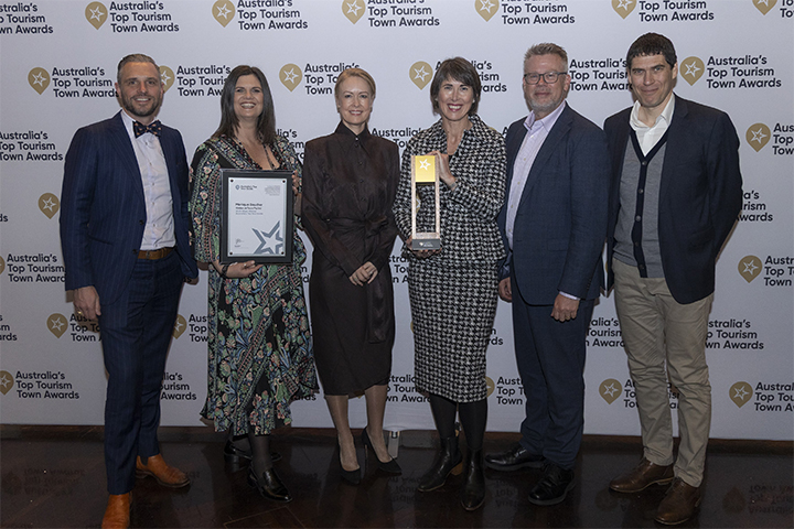 Six people stand together at Australias Top Tourism Town Awards, smiling for the camera. One person holds a framed certificate, another displays a trophy. A branded event backdrop is behind them.