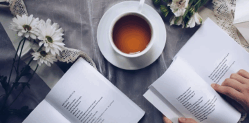 A cup of tea sits on a saucer near open books of poetry and a bouquet of white flowers, all arranged on a table with a lace tablecloth. Two hands are holding and reading the books.