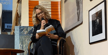 A woman with blonde hair and glasses sits in a chair, reading from a book and speaking into a microphone. She is inside a room decorated with photographs and posters on the walls.