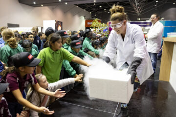 A scientist in a lab coat and goggles handles a smoking box, possibly with dry ice, as a group of excited children in school uniforms reach out toward the mist during a science demonstration.