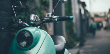 A close-up of a teal scooter parked on a street next to a wall with greenery; the background is blurred, showing an alleyway and a person walking in the distance.