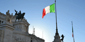 An Italian flag waves in the wind on a tall flagpole, with a historic white marble building and equestrian statue under a clear blue sky in the background.