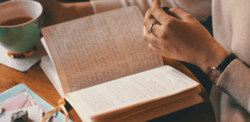A person wearing a ring is sitting at a table, holding an open journal filled with handwritten notes. There is a cup of tea and some colorful papers nearby.