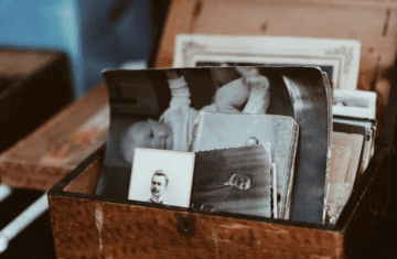 A wooden box filled with old black-and-white photographs, some showing portraits of people, sits open on a table. The edges of the photos appear worn with age.