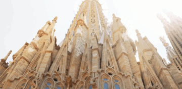A low-angle view of the ornate spires and detailed stonework of the Sagrada Família basilica in Barcelona, with sunlight illuminating the structure from behind.