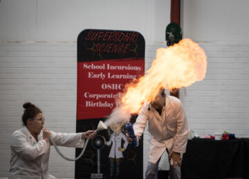 Two people in lab coats perform a science experiment, creating a large fireball. They are in front of a banner reading Supersonic Science and a white brick wall. Scientific props are on a table nearby.