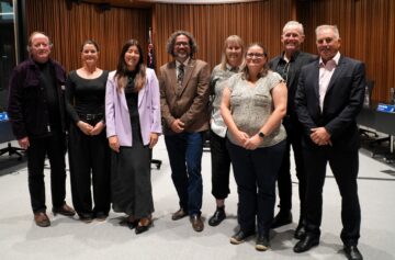 Eight people stand in a row, smiling, in a formal indoor setting with an Australian flag and wood-paneled walls in the background. Several are dressed in business attire.