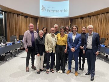 A group of ten people stand smiling in a council chamber with a “City of Fremantle” logo projected behind them. They are dressed in business and casual attire, standing in front of a semicircle of desks and chairs.