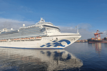 A large white cruise ship named Crown Princess sails near a port with shipping containers and a red crane, reflecting on calm blue water under a clear sky.