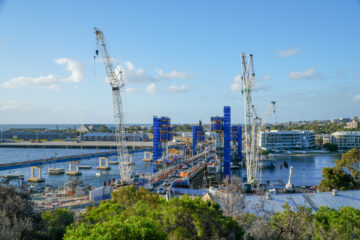Construction site with several large cranes and scaffolding over a river, building a new bridge. Modern buildings and trees are visible nearby under a bright blue sky with scattered clouds.