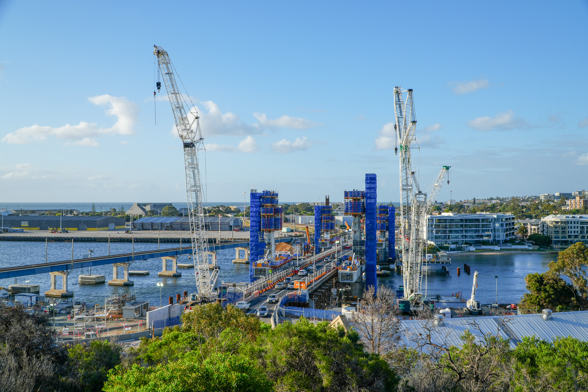 Construction site with several large cranes and scaffolding over a river, building a new bridge. Modern buildings and trees are visible nearby under a bright blue sky with scattered clouds.