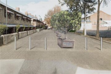A paved street lined with rectangular planters containing leafy trees, bordered by buildings on the left and a fence and building on the right, on a partly cloudy day.