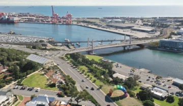 Aerial view of a large bridge crossing a river, with industrial port cranes, warehouses, and docks in the background; cars and green spaces in the foreground near a colorful art installation.