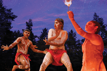 Three Indigenous Australian performers with traditional body paint and red clothing dance outdoors at dusk, with one person holding a flaming object. Tall trees and a starry purple sky are visible in the background.