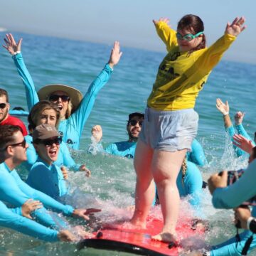 A young woman in a yellow shirt stands on a surfboard, smiling as a group of people in blue shirts cheer her on in the ocean. The group is excited and supportive, with arms raised in celebration.
