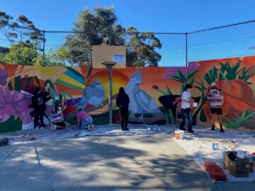 A group of people paint a colorful mural with flowers and birds on a wall at an outdoor basketball court. Paint cans and supplies are scattered on tarps covering the ground. Trees and a blue sky are visible in the background.