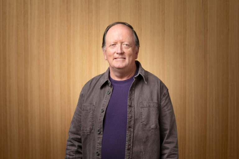 A middle-aged man with short hair stands smiling in front of a wooden panel background. He is wearing a purple shirt and a gray button-up jacket.
