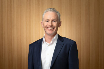 A smiling man with short gray hair, wearing a white shirt and dark blue blazer, stands in front of a light wooden background.