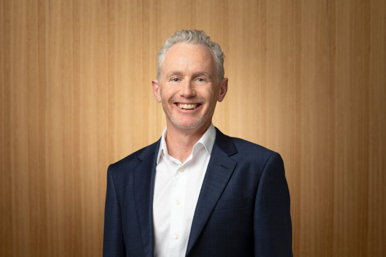 A smiling man with short gray hair, wearing a white shirt and dark blue blazer, stands in front of a light wooden background.