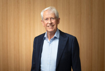 An older man with white hair, wearing a navy blue blazer and light blue shirt, stands smiling in front of a light wood-paneled background.