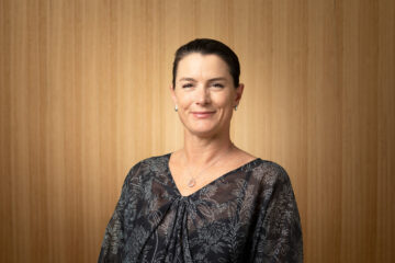 A woman with dark hair pulled back, wearing a dark floral blouse and a necklace, stands smiling in front of a light wooden panel background.